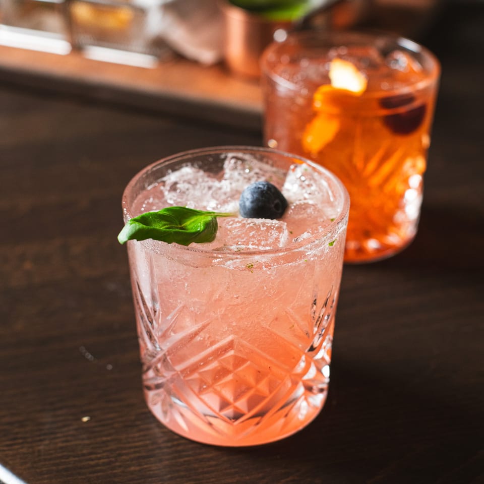 Two colorful cocktails sit on a dark wooden table, including a pink drink garnished with mint and a blueberry.
