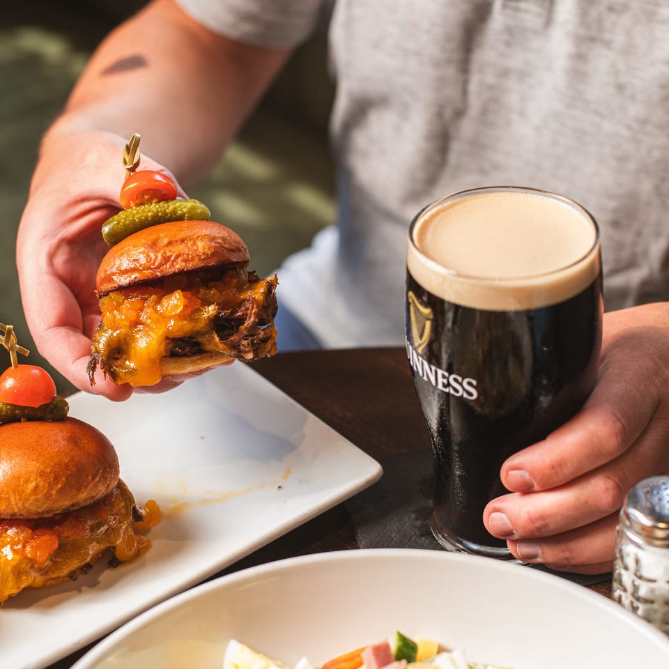 Diner holds a cheeseburger topped with tomato and pickle beside a pint of Guinness, with additional burgers and salad on the table.