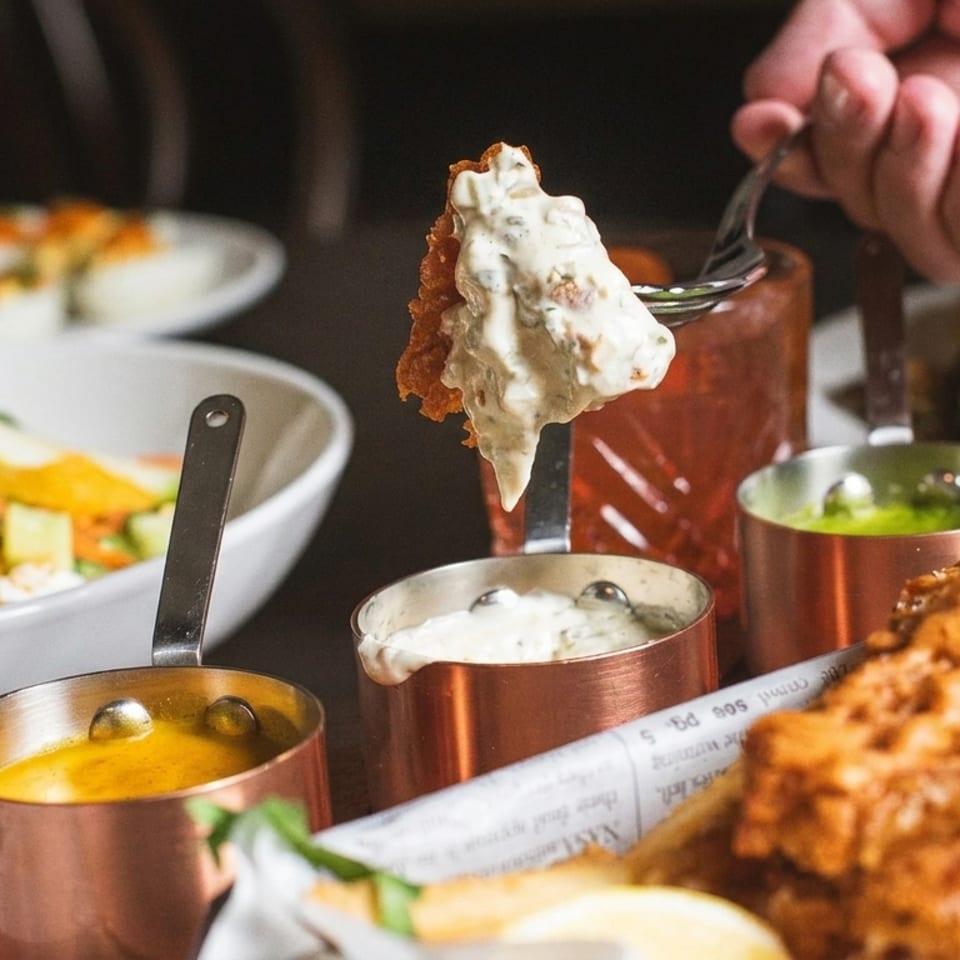 Close-up of fried chicken dipped in creamy sauce, surrounded by colorful dishes, dipping cups, and a burger at a restaurant table.