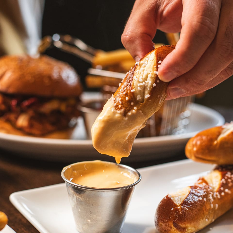 Hand dips a salt-topped pretzel into warm cheese sauce beside additional pretzels on a serving tray.