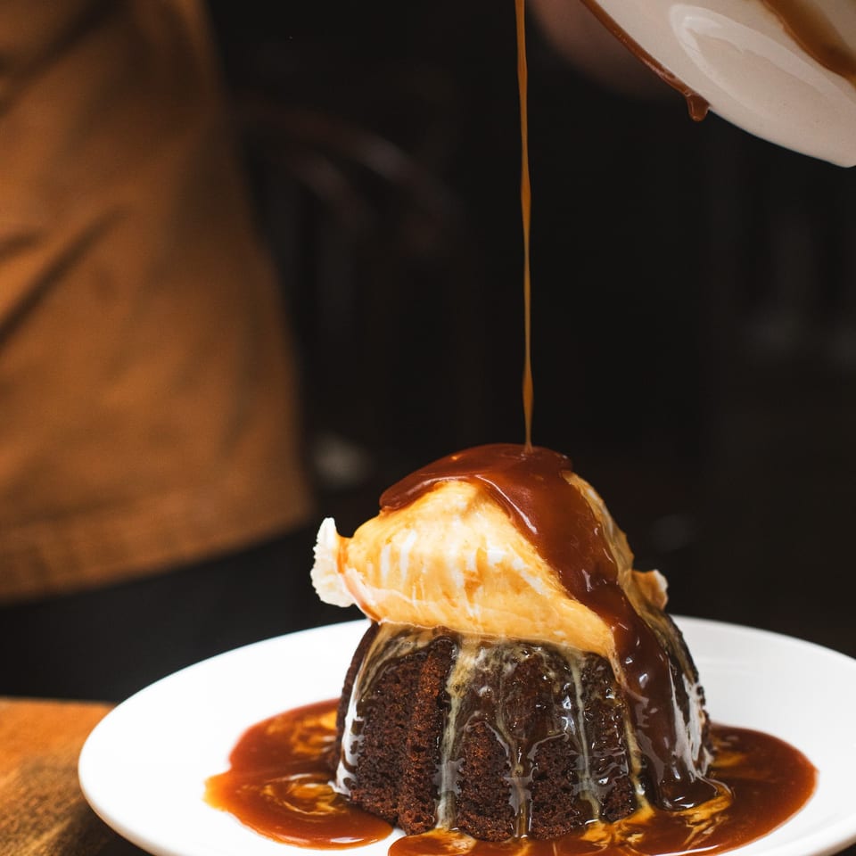 Chocolate cake topped with vanilla ice cream and glossy chocolate sauce being poured onto a white plate.