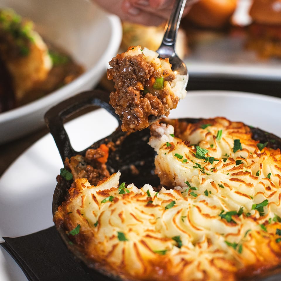 Fork lifts a bite of savory meat and vegetables from a skillet topped with golden mashed potatoes and herbs.