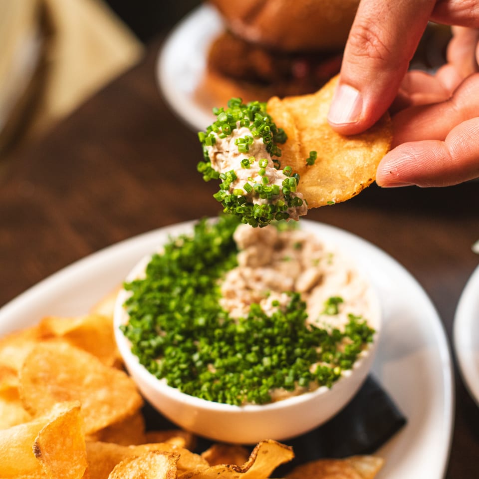 Hand dips a crisp tortilla chip into creamy dip topped with chopped green onions, surrounded by more chips.
