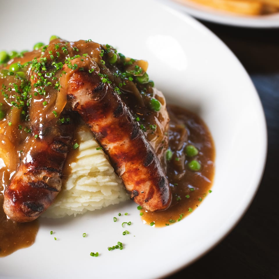Grilled sausage and roasted meat with mashed potatoes, brown gravy, and chopped chives on a white plate.